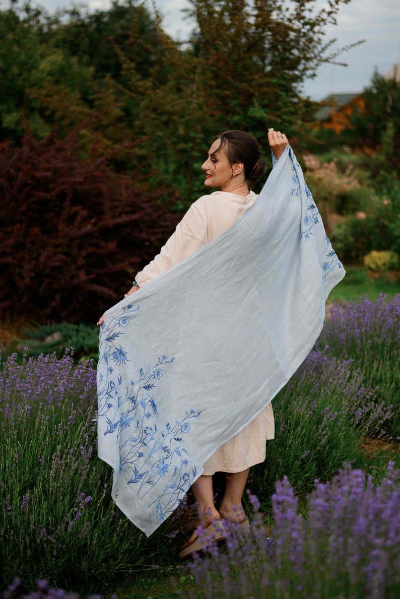 K&ouml;nnte beinhalten: Eine Frau in einem hellbeigen Kleid h&auml;lt einen hellblauen Schal mit Blumenmuster. Der Schal hat ein blaues Blumenmuster am unteren Rand. Die Frau steht auf einem Lavendelfeld.