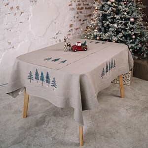 May include: A beige linen tablecloth with embroidered evergreen trees and falling snow. A red toy truck carrying a small green Christmas tree and a silver reindeer figurine are on the table. A decorated Christmas tree is in the background.