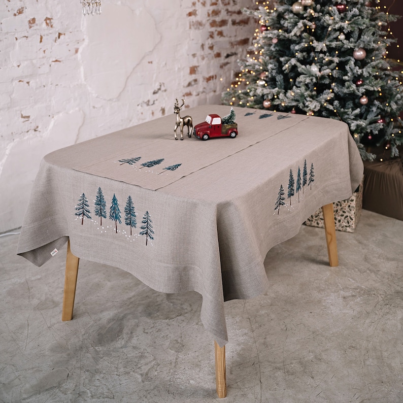 May include: A beige linen tablecloth with embroidered evergreen trees and falling snow. A red toy truck carrying a small green Christmas tree and a silver reindeer figurine are on the table. A decorated Christmas tree is in the background.