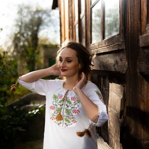 May include: A woman wearing a white embroidered blouse with a floral design and red berries. She is standing in front of a wooden building with a large window.