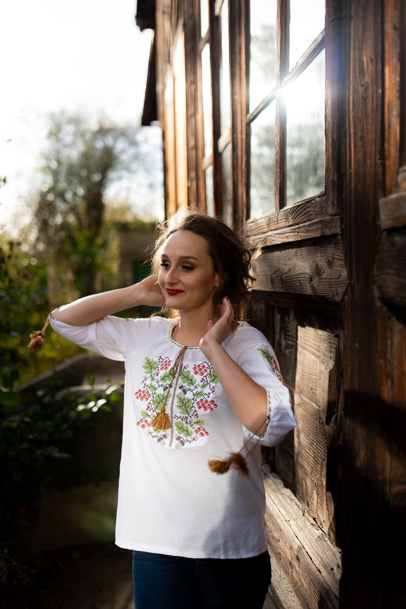 May include: A woman wearing a white embroidered blouse with a floral design and red berries. She is standing in front of a wooden building with a large window.