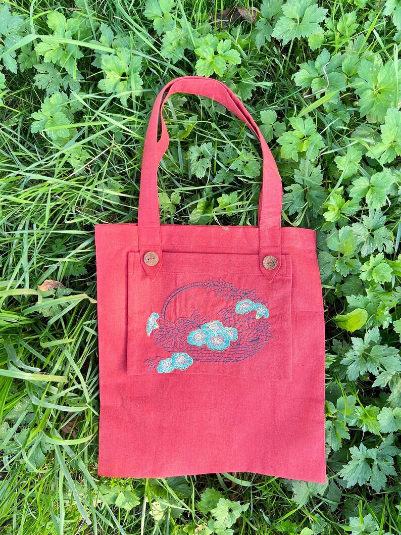 Op de afbeelding: Een rode tote bag met een geborduurd bloemenontwerp op de voorzak. De tas heeft twee bruine knopen aan de voorkant.