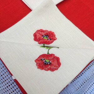 May include: Two white linen napkins with red embroidered poppies. The napkins are folded and layered on top of each other.