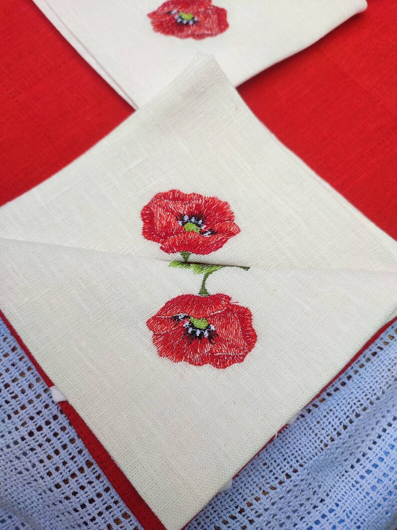 May include: Two white linen napkins with red embroidered poppies. The napkins are folded and layered on top of each other.