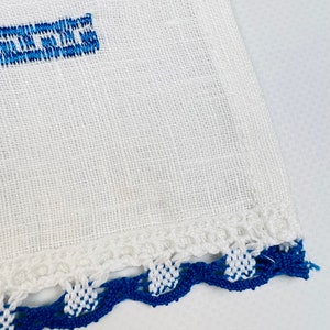 Peut inclure: Tissu de lin blanc avec texte brod&eacute; bleu et bordure crochet&eacute;e d&eacute;corative. La bordure pr&eacute;sente un motif festonn&eacute; avec des points de crochet blancs et bleus altern&eacute;s, ajoutant une touche artisanale.