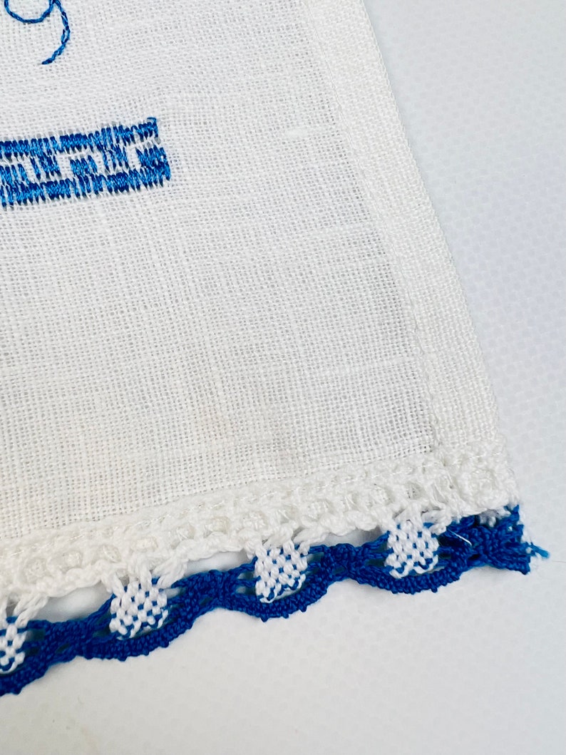 Peut inclure: Tissu de lin blanc avec texte brod&eacute; bleu et bordure crochet&eacute;e d&eacute;corative. La bordure pr&eacute;sente un motif festonn&eacute; avec des points de crochet blancs et bleus altern&eacute;s, ajoutant une touche artisanale.