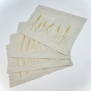 Op de afbeelding: Vier rechthoekige linnen placemats met geborduurde tarwearen-ontwerpen. De tarwe is afgebeeld in goud en gebroken wit op een neutrale beige achtergrond. Elke placemat meet ongeveer 30 cm x 40 cm.