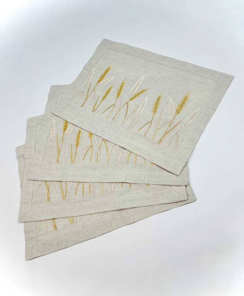 Op de afbeelding: Vier rechthoekige linnen placemats met geborduurde tarwearen-ontwerpen. De tarwe is afgebeeld in goud en gebroken wit op een neutrale beige achtergrond. Elke placemat meet ongeveer 30 cm x 40 cm.
