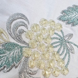 以下が含まれることがあります： 詳細な刺繍が施された白い生地のクローズアップ。デザインは、クリーム色のふわふわの花の要素、銀色の渦巻き、青緑色の葉のクラスターを特徴としています。刺繍は複雑にステッチされ、テクスチャード加工された表面を作り出しています。