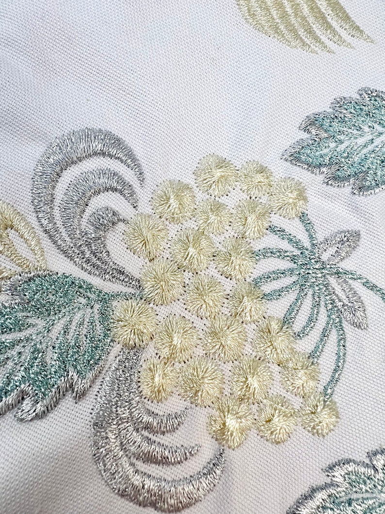 以下が含まれることがあります： 詳細な刺繍が施された白い生地のクローズアップ。デザインは、クリーム色のふわふわの花の要素、銀色の渦巻き、青緑色の葉のクラスターを特徴としています。刺繍は複雑にステッチされ、テクスチャード加工された表面を作り出しています。