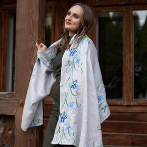 May include: A light gray shawl with embroidered blue floral designs and green stems. The shawl is draped over a dark green dress. The background features a wooden structure with windows.
