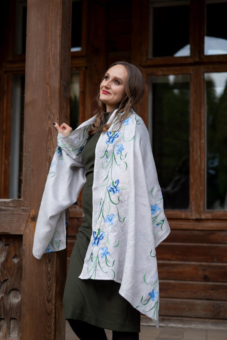 May include: A light gray shawl with embroidered blue floral designs and green stems. The shawl is draped over a dark green dress. The background features a wooden structure with windows.
