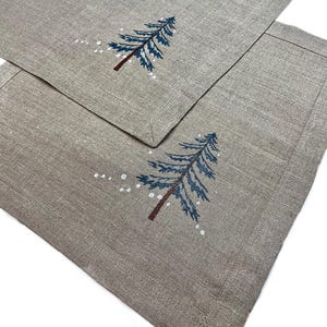 May include: Two rectangular placemats in a neutral beige color. Each placemat features an embroidered blue Christmas tree with a brown trunk and white snow accents. The placemats have a stitched border.