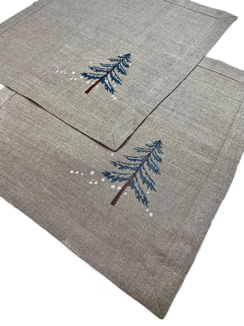 May include: Two rectangular placemats in a neutral beige color. Each placemat features an embroidered blue Christmas tree with a brown trunk and white snow accents. The placemats have a stitched border.