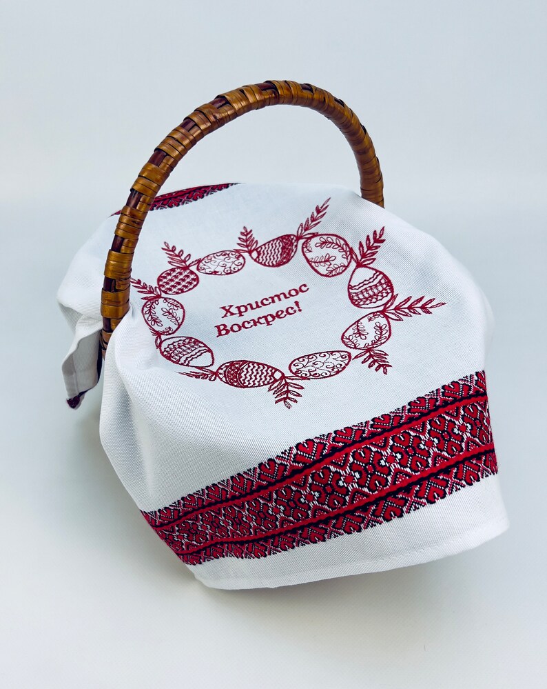 May include: White fabric Easter basket with a woven handle. The fabric has a red embroidered border and a wreath of red embroidered eggs with the text "Христос Воскрес!" in the center.