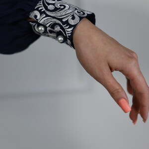 May include: A navy blue long-sleeved shirt with a white embroidered cuff. The cuff features a floral design and two pearl-like buttons.