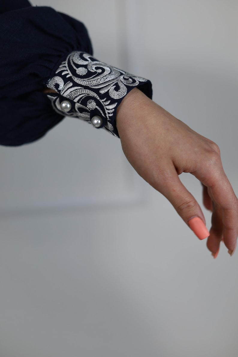 May include: A navy blue long-sleeved shirt with a white embroidered cuff. The cuff features a floral design and two pearl-like buttons.