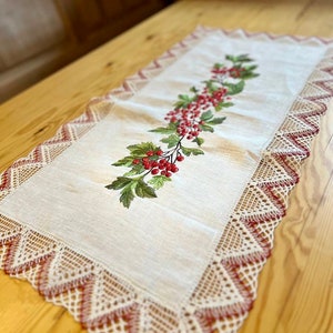 May include: A white linen table runner with a red berry and green leaf embroidery design. The runner has a delicate lace border with red accents.