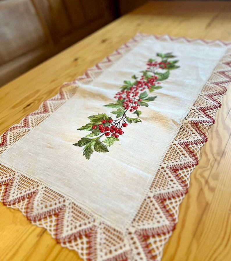 May include: A white linen table runner with a red berry and green leaf embroidery design. The runner has a delicate lace border with red accents.