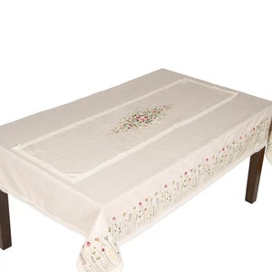 May include: A rectangular beige tablecloth with floral embroidery and lace trim. The embroidery features a cluster of pink, red, and green flowers in the center and along the edges. The tablecloth is on a dark brown table.