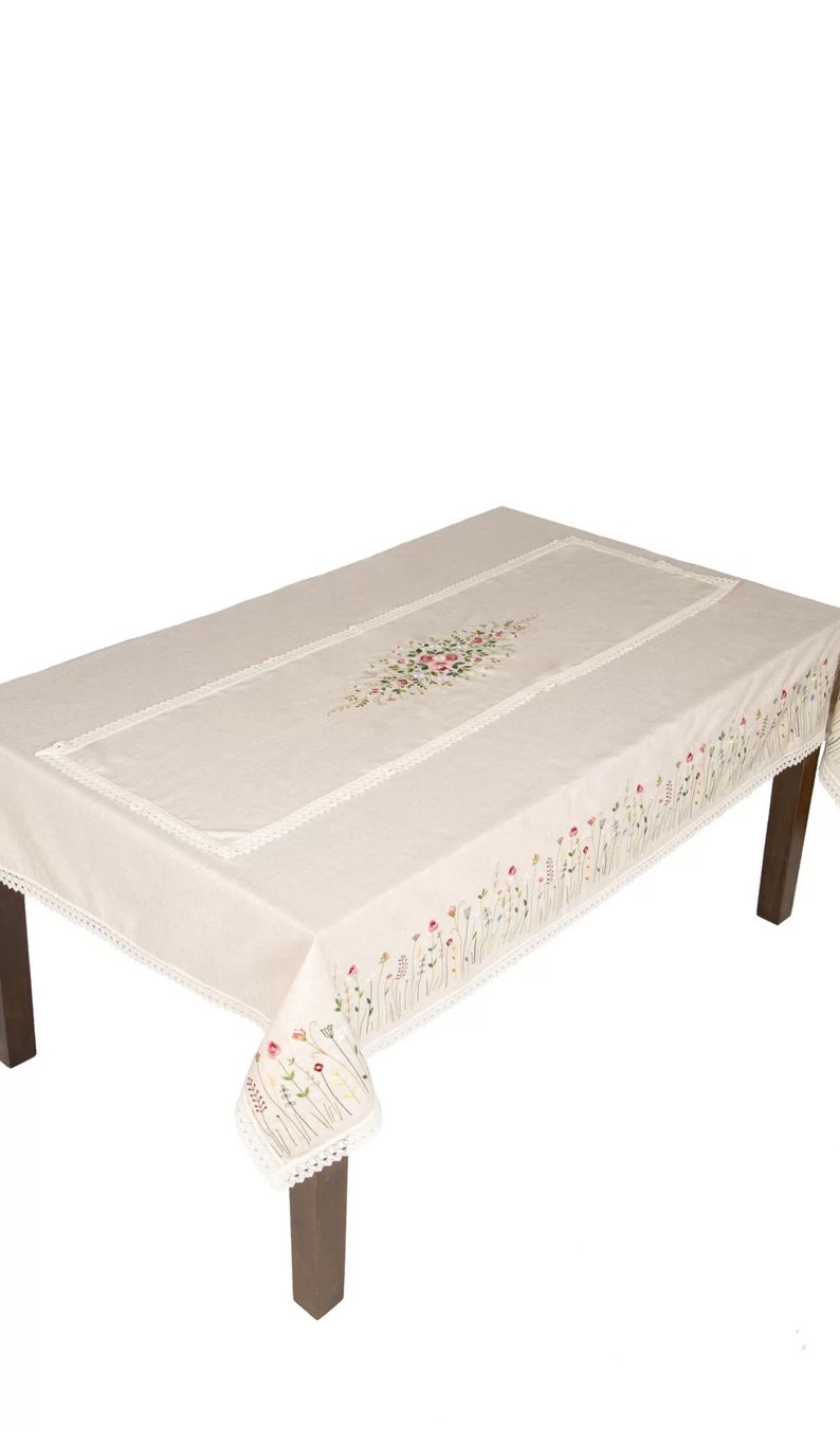 May include: A rectangular beige tablecloth with floral embroidery and lace trim. The embroidery features a cluster of pink, red, and green flowers in the center and along the edges. The tablecloth is on a dark brown table.