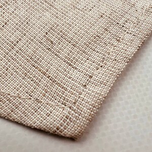 May include: Close-up of a beige fabric with a woven texture. The fabric has a stitched edge.