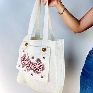 Op de afbeelding: Een witte linnen tote bag met een rood en zwart geborduurd ontwerp. De tas heeft een klep met een houten knoopsluiting en een kleiner vakje aan de voorkant.
