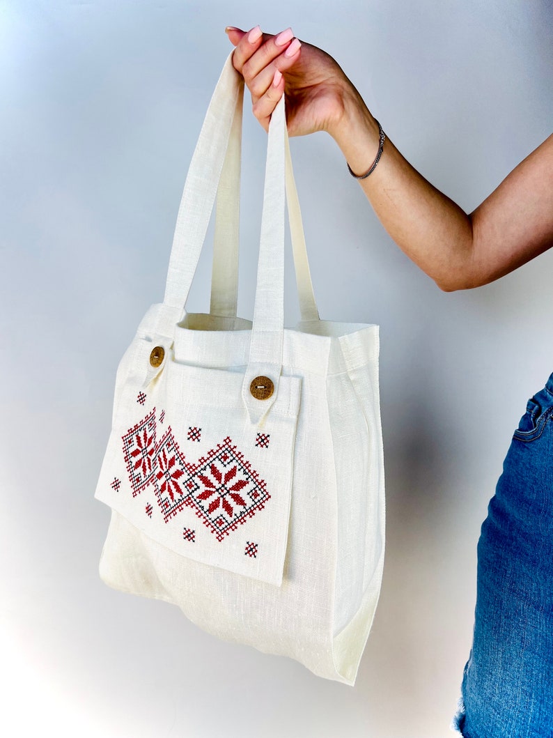 Op de afbeelding: Een witte linnen tote bag met een rood en zwart geborduurd ontwerp. De tas heeft een klep met een houten knoopsluiting en een kleiner vakje aan de voorkant.
