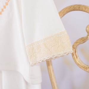 May include: White short-sleeved shirt with a delicate lace trim around the sleeves. The lace is a light beige color.