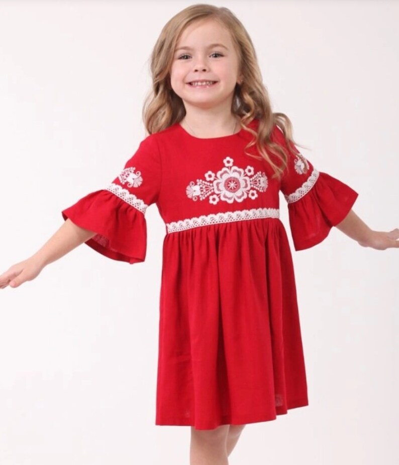 K&ouml;nnte beinhalten: Ein rotes Leinenkleid mit wei&szlig;en gestickten Blumen und Spitzenbesatz. Das Kleid hat einen runden Ausschnitt und 3/4 &Auml;rmel.