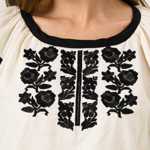 May include: Close-up of a cream-coloured top with black floral embroidery. The top features a black neckline and a vertical black embroidered design down the centre. The sleeves are puffed and the fabric appears to be linen or a similar material.