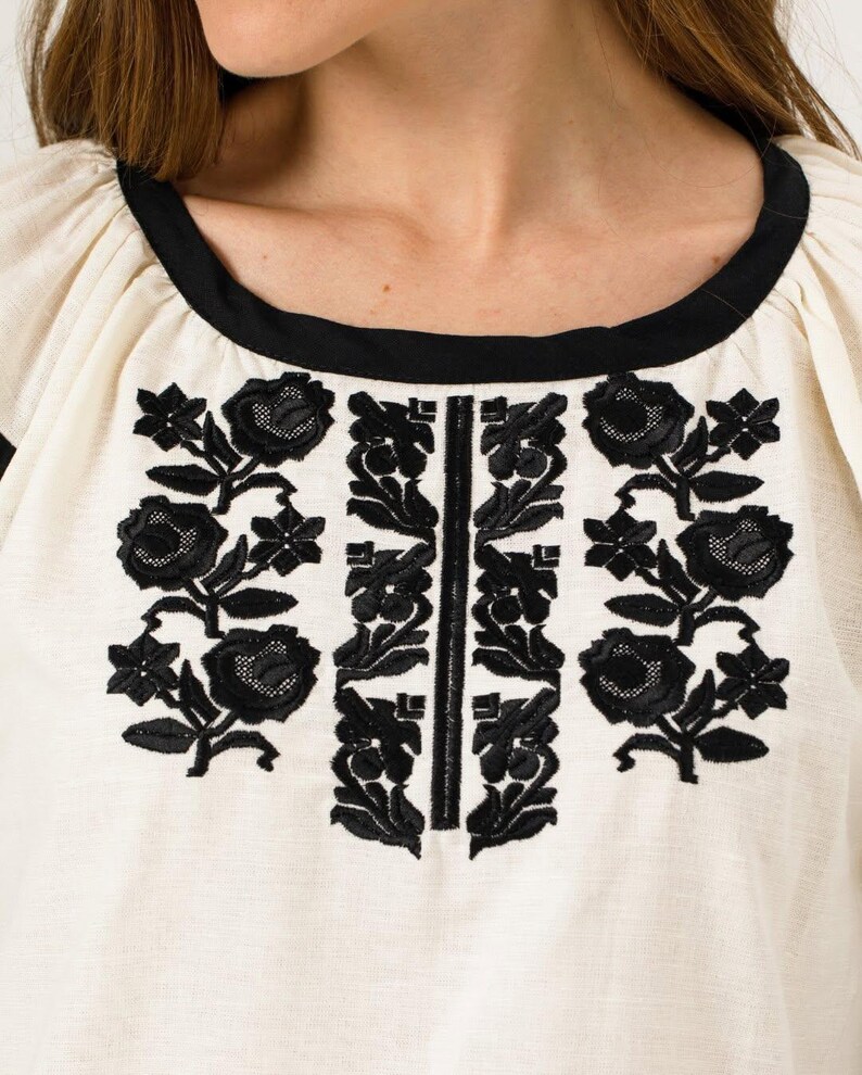 May include: Close-up of a cream-coloured top with black floral embroidery. The top features a black neckline and a vertical black embroidered design down the centre. The sleeves are puffed and the fabric appears to be linen or a similar material.