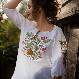 May include: A white embroidered blouse with a floral design featuring red berries and green leaves. The blouse has a tassel detail at the neckline.
