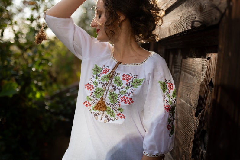 May include: A white embroidered blouse with a floral design featuring red berries and green leaves. The blouse has a tassel detail at the neckline.