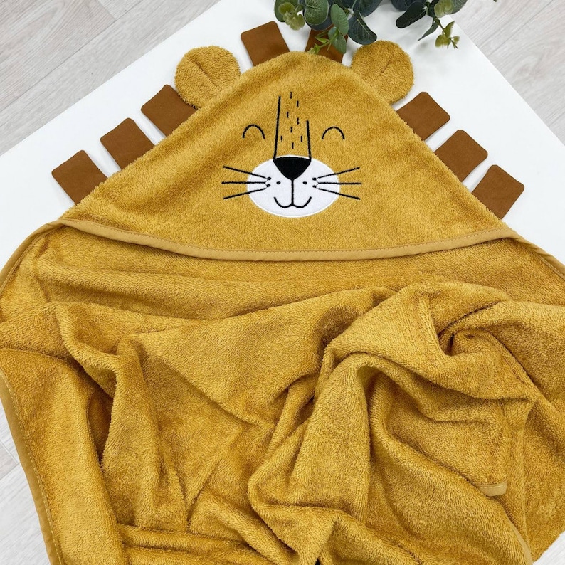 May include: A mustard yellow hooded towel with a lion face embroidered on the hood. The lion has black eyes, a black nose, and a white mouth with a smile.