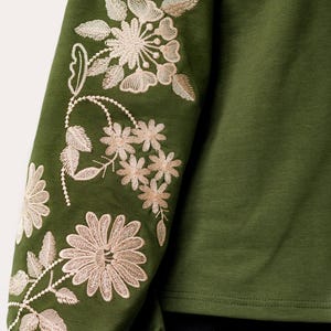 以下が含まれることがあります： 袖に刺繍された花柄のディテールが施された緑色の長袖シャツ。花は薄いベージュ色です。