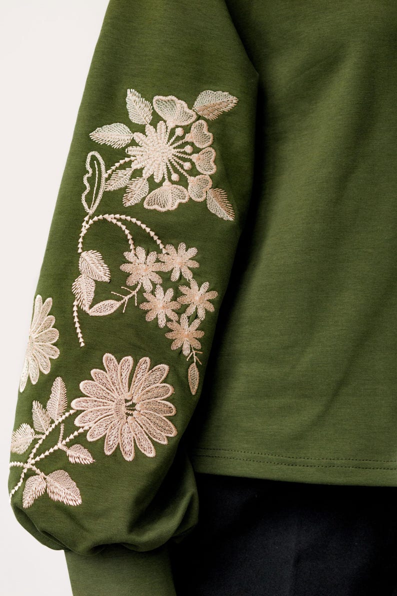 以下が含まれることがあります： 袖に刺繍された花柄のディテールが施された緑色の長袖シャツ。花は薄いベージュ色です。