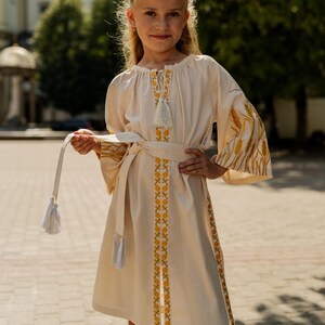 K&ouml;nnte beinhalten: Cremefarbenes Kleid mit langen &Auml;rmeln und passendem G&uuml;rtel. Das Kleid hat gelbe und goldene Stickereien am Ausschnitt, an den &Auml;rmeln und am Saum. Die &Auml;rmel haben ein weizen&auml;hnliches Design. Der G&uuml;rtel hat Quasten.