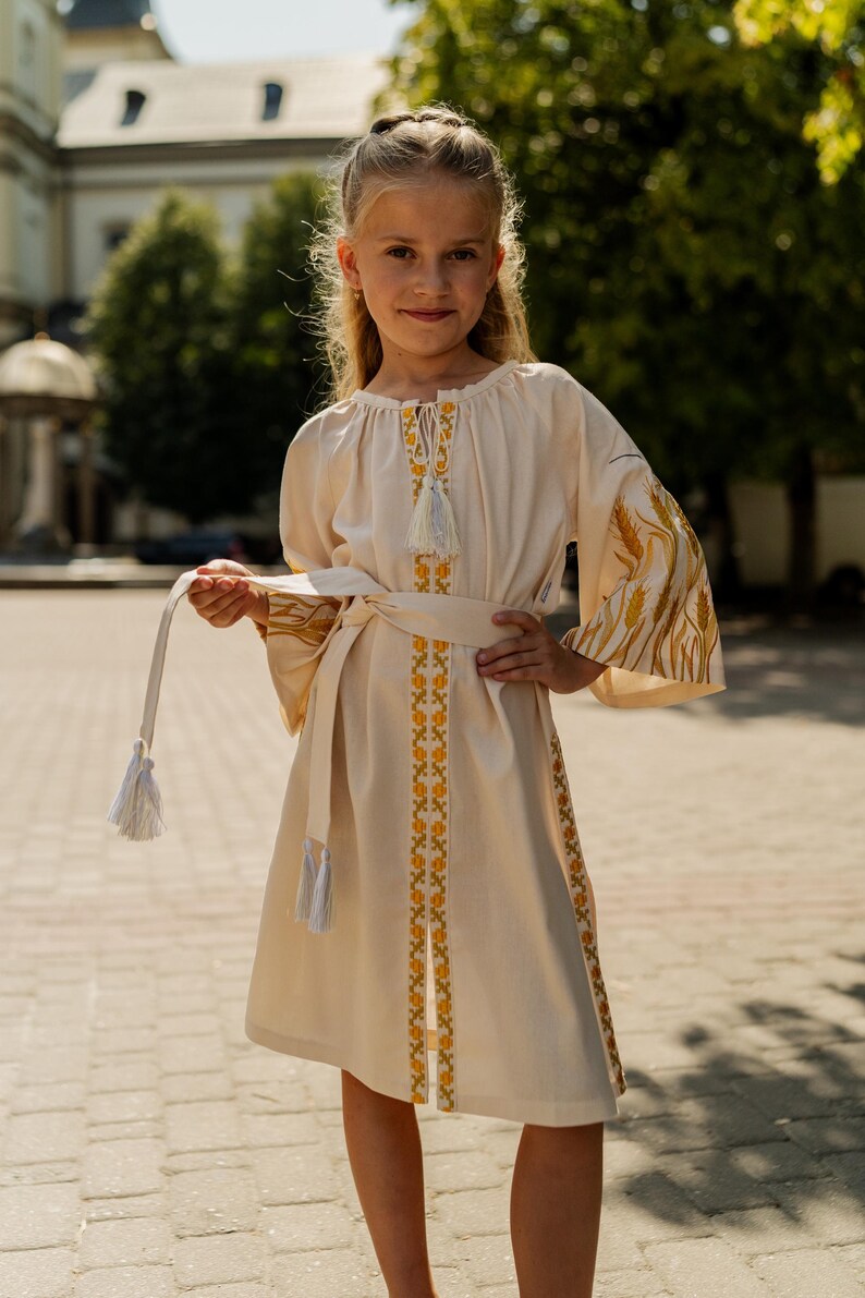 K&ouml;nnte beinhalten: Cremefarbenes Kleid mit langen &Auml;rmeln und passendem G&uuml;rtel. Das Kleid hat gelbe und goldene Stickereien am Ausschnitt, an den &Auml;rmeln und am Saum. Die &Auml;rmel haben ein weizen&auml;hnliches Design. Der G&uuml;rtel hat Quasten.