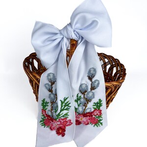 May include: A brown wicker basket with a light blue satin bow and a white fabric scarf. The scarf is embroidered with a floral design featuring red flowers, green leaves, and gray buds.