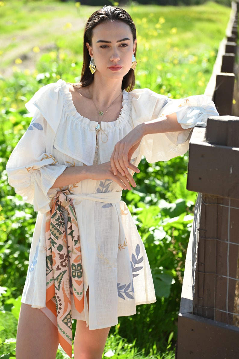 May include: A white linen dress with a ruffled neckline and a floral print. The dress has a tie belt and a scarf with a geometric pattern.