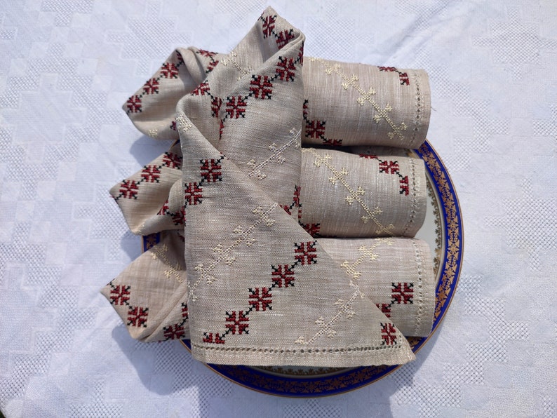 May include: A set of beige linen napkins with embroidered red and black cross patterns, arranged on a blue-rimmed plate. The napkins are folded and rolled, showcasing the detailed needlework. Ideal for home decor.