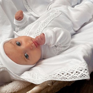 Op de afbeelding: Een witte baby-outfit met kantversiering, gedragen door een baby die op een witte deken met kantversiering ligt. De baby draagt een witte muts en heeft blauwe ogen.