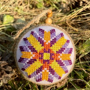 May include: A round, white ornament with a purple, orange, and yellow geometric design. The ornament is made of fabric and has a brown twine string attached.