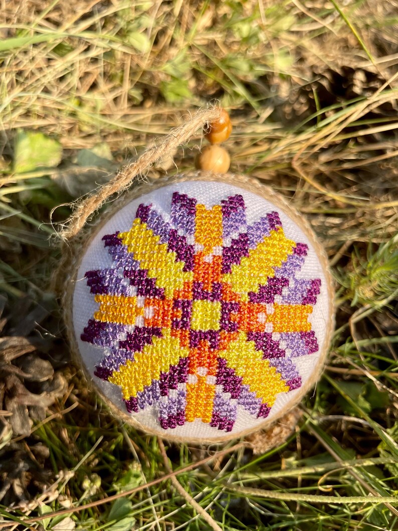 May include: A round, white ornament with a purple, orange, and yellow geometric design. The ornament is made of fabric and has a brown twine string attached.