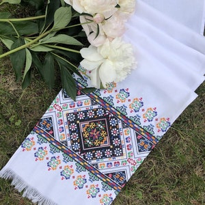 May include: White embroidered table runner with a colorful geometric pattern and fringe trim. The runner is laid out on green grass with white peonies in the background.