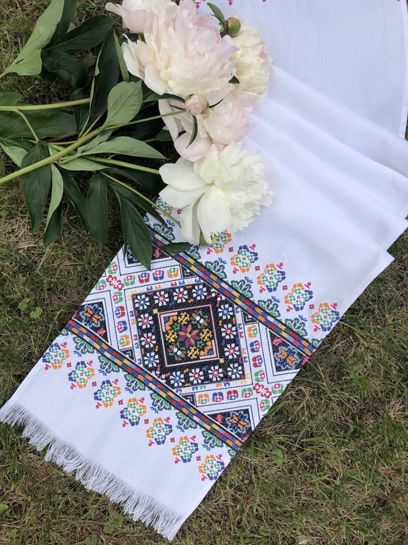 May include: White embroidered table runner with a colorful geometric pattern and fringe trim. The runner is laid out on green grass with white peonies in the background.