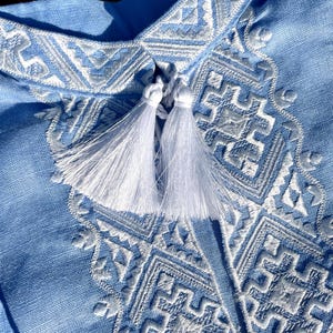 May include: Close-up of a light blue shirt with intricate white embroidery. The shirt features a detailed geometric pattern around the collar and chest, with two white tassels. The fabric appears to be linen or a similar lightweight material.