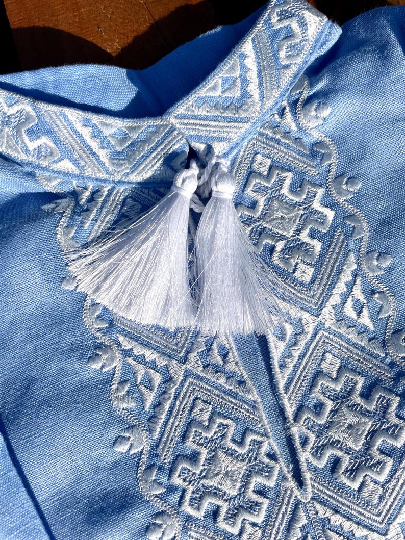 May include: Close-up of a light blue shirt with intricate white embroidery. The shirt features a detailed geometric pattern around the collar and chest, with two white tassels. The fabric appears to be linen or a similar lightweight material.