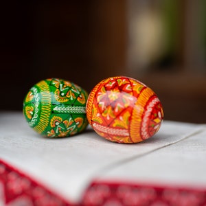 K&ouml;nnte beinhalten: Zwei dekorierte Ostereier. Ein Ei ist gr&uuml;n mit gelben und wei&szlig;en Blumenmustern. Das andere Ei ist rot und orange mit aufwendigen Designs. Sie liegen auf einem wei&szlig;en Tuch mit einem rot-wei&szlig; gemusterten Rand.